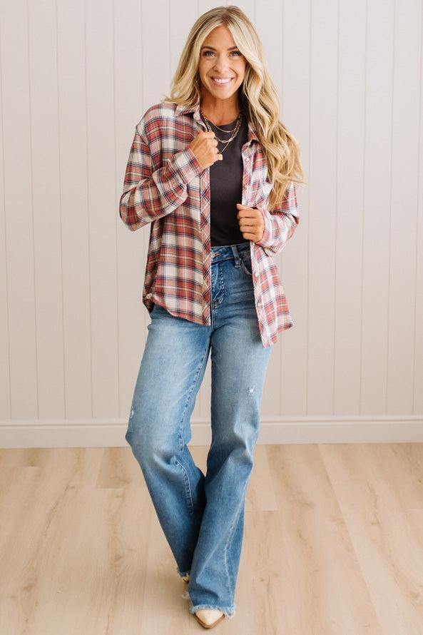 A woman modeling a red and white plaid button-down shirt over a black top and blue jeans, standing in front of a white paneled wall and light wood floor.