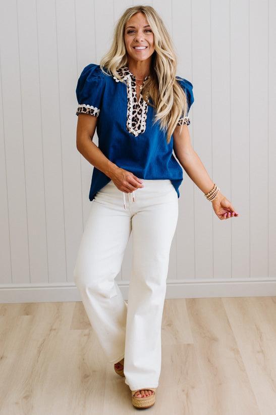 Woman wearing a blue top with lace details and white pants against a light wooden floor and wall background.