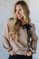 Woman wearing a beige sweater with black ribbons against a white brick wall
