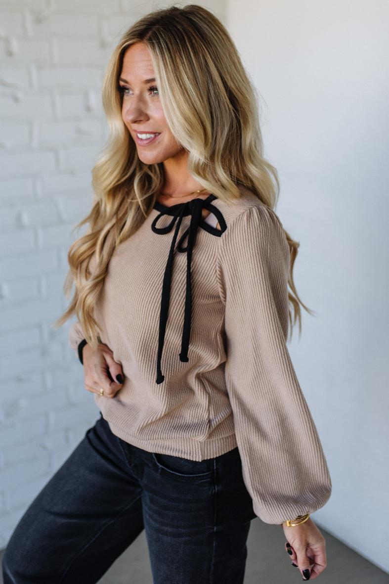 Woman wearing a beige blouse with black bow details against a white brick wall.