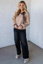 Woman wearing a beige blouse with black bow details and dark jeans against a white brick wall.