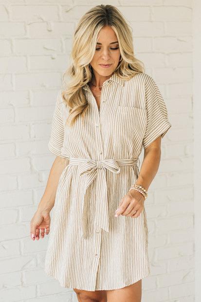 Blonde woman wearing a knee length striped shirt dress with button front and matching sash at waist.