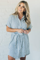 Blonde woman wearing a knee length striped shirt dress with button front and matching sash at waist.