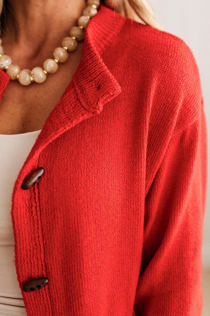 Blonde woman in dark jeans and a bold red-orange cardigan with black button toggles down with front and side pockets.