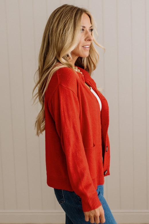 Blonde woman in dark jeans and a bold red-orange cardigan with black button toggles down with front and side pockets.