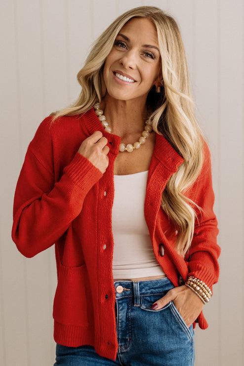 Blonde woman in dark jeans and a bold red-orange cardigan with black button toggles down with front and side pockets.
