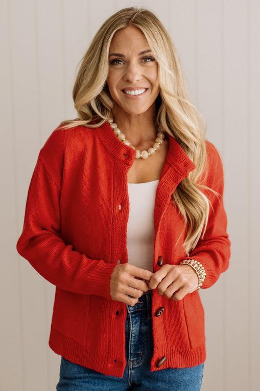 Blonde woman in dark jeans and a bold red-orange cardigan with black button toggles down with front and side pockets.
