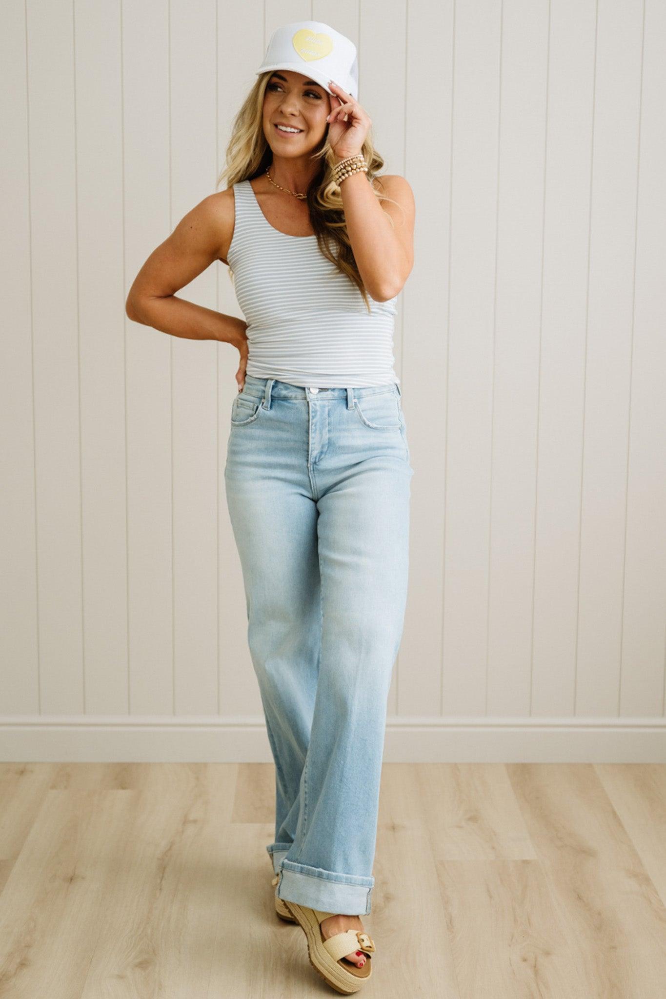 Blonde woman wearing long length light wash denim jeans with a cuff at the ankle. Wearing with a striped tank and white trucker hat.