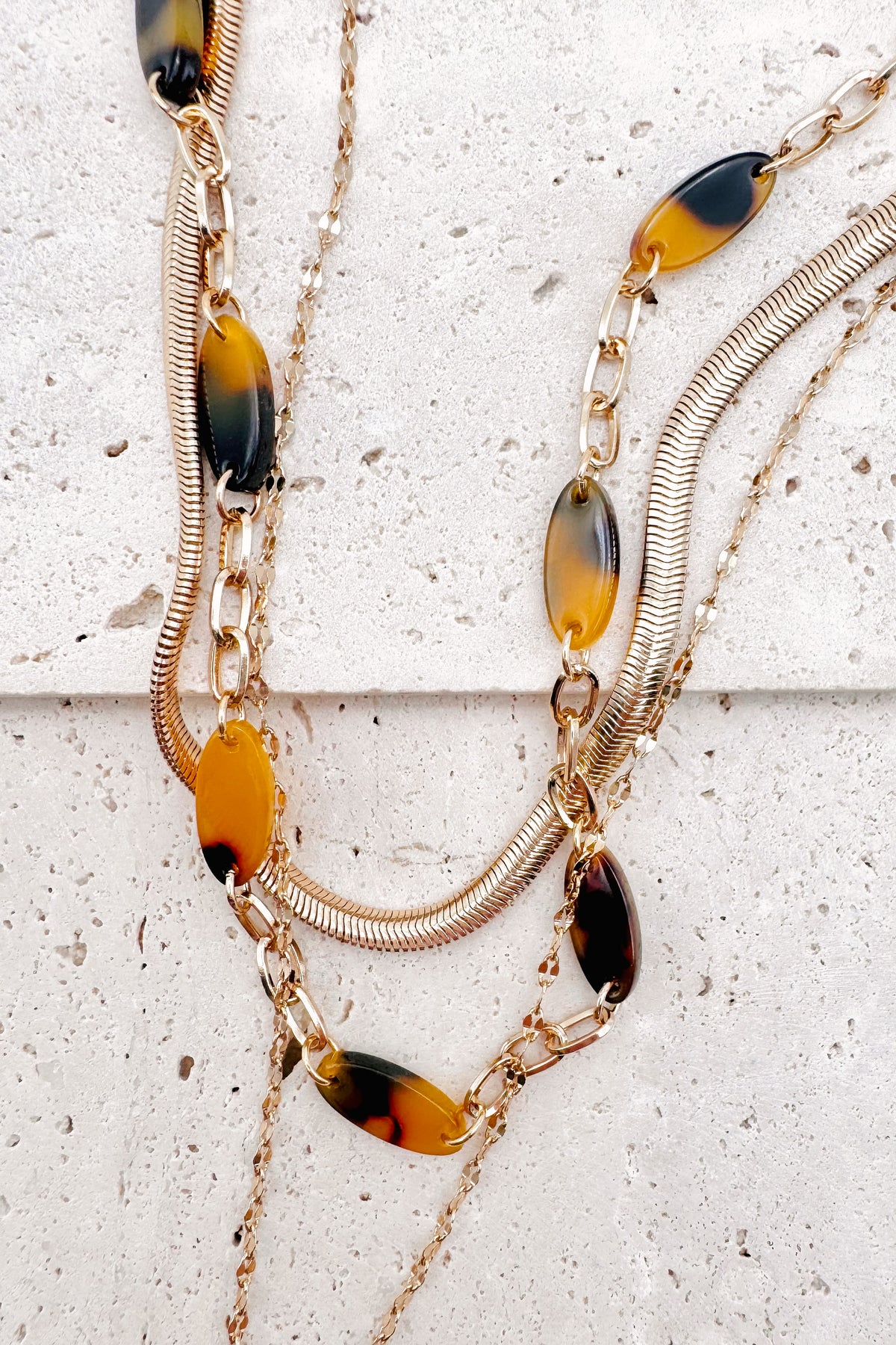 A refined layered necklace featuring a sleek gold herringbone chain paired with a delicate link strand accented by warm tortoiseshell-style acetate beads. A petite heart coin pendant adds a soft feminine touch, creating an elegant balance between modern texture and timeless shine. The adjustable length allows the layers to sit perfectly along the neckline for effortless styling.