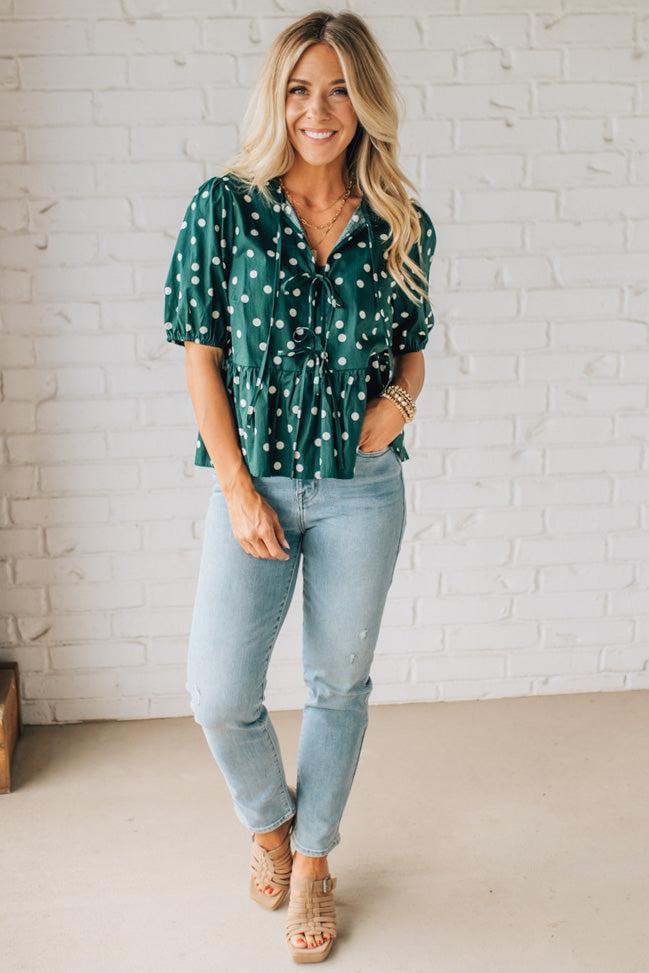 Woman wearing a green polka dot blouse and light blue jeans against a white brick wall.