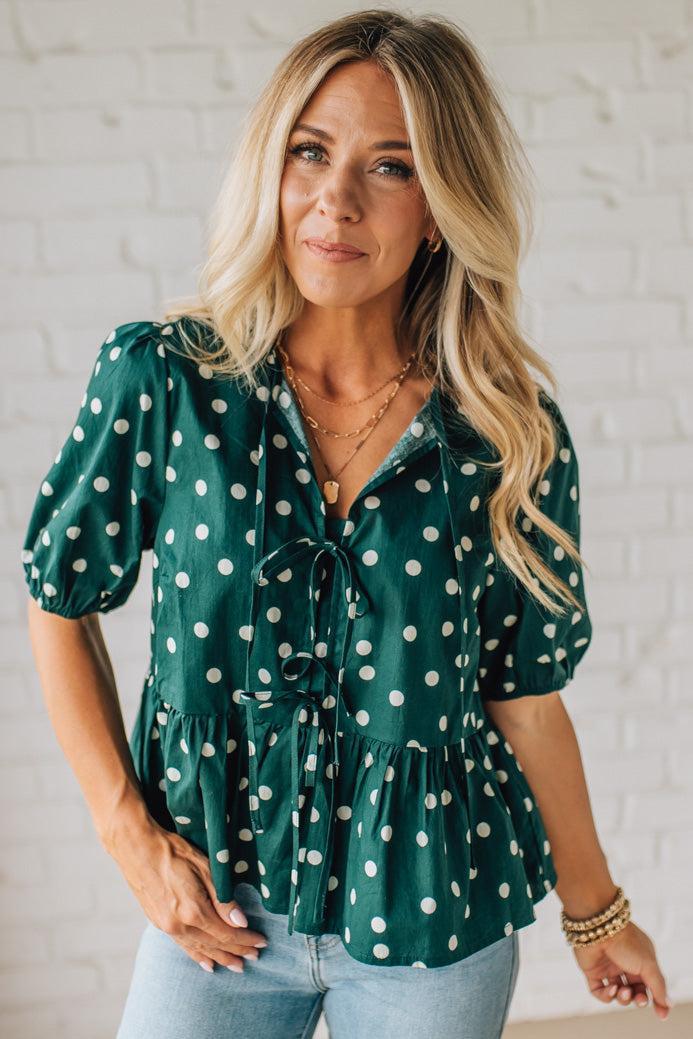 Woman wearing a green polka dot blouse against a white brick wall.