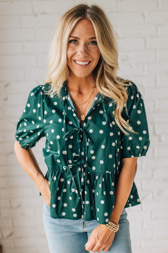 Woman wearing a green polka dot blouse against a white brick wall.