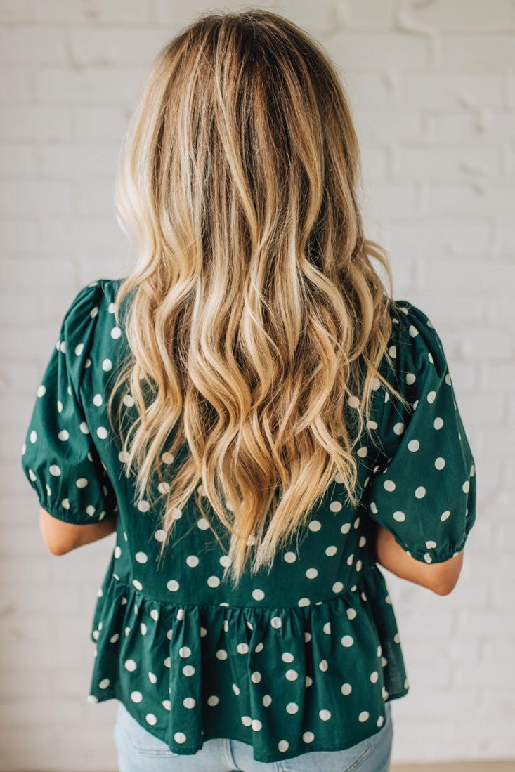 Person wearing a green polka dot blouse with long blonde hair, standing against a light brick wall.
