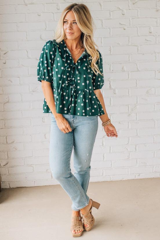 Woman wearing a green polka dot blouse and light blue jeans against a white brick wall.
