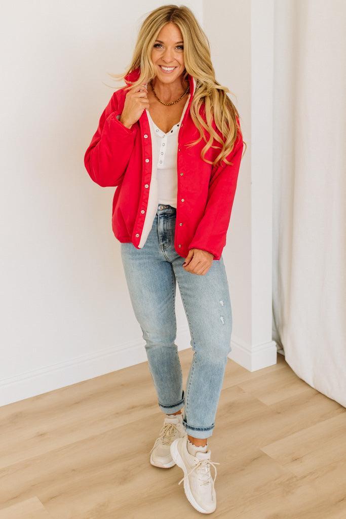 Woman wearing a red cardigan, white shirt, blue jeans, and white sneakers in a room with a white wall and wooden floor.