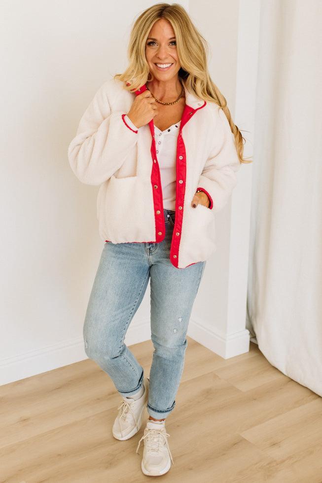 Woman wearing a white cardigan with red trim, white shirt, blue jeans, and white sneakers indoors.
