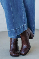 Brown crocodile-patterned boots worn with blue jeans on a light gray floor.
