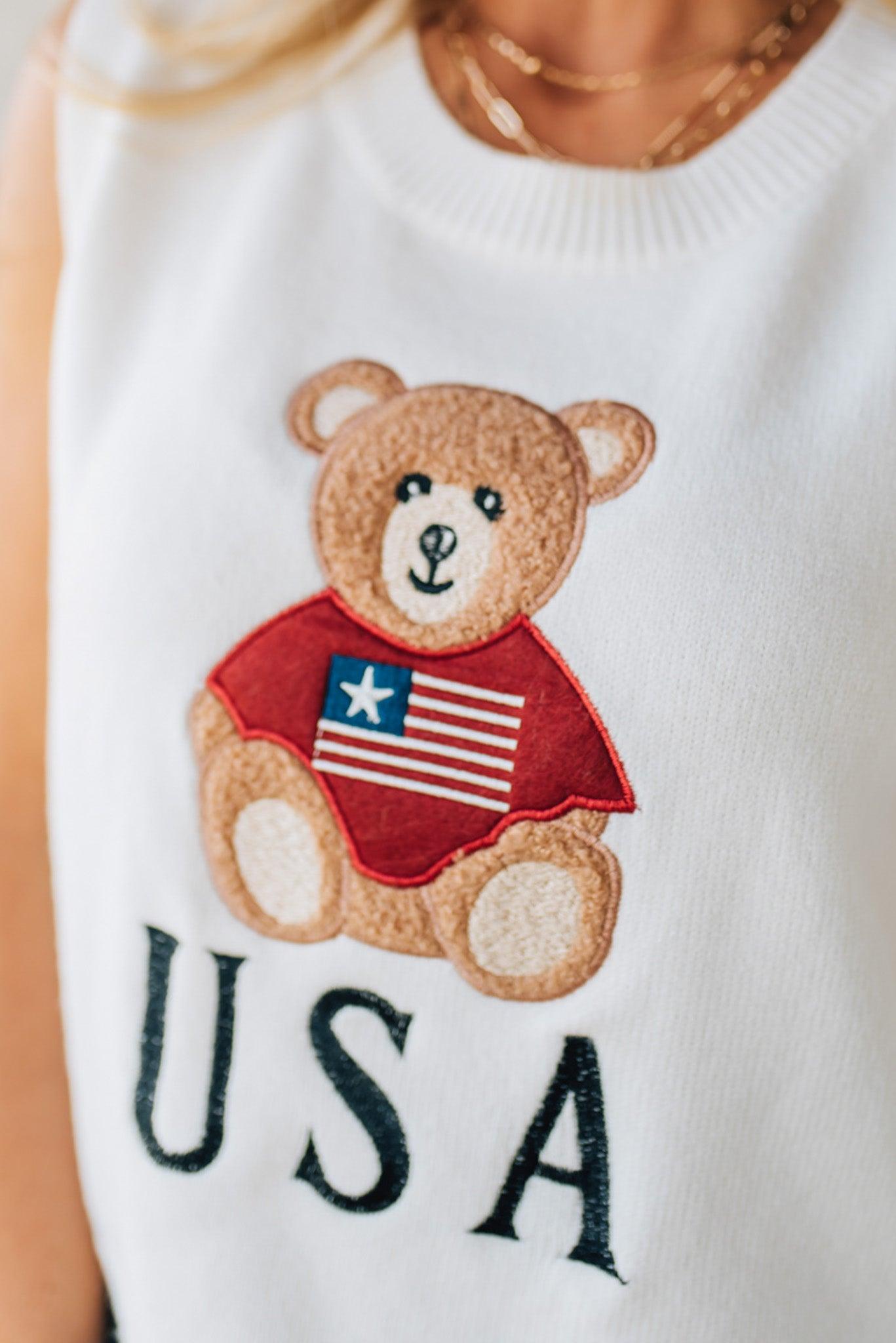 BLONDE WOMAN IN DENIM SHORTS WEARING A TEDDY BEAR WEARING AN AMERICAN FLAG WITH USA EMBROIDERY LETTERING FOURTH OF JULY SWEATER TANK TOP.