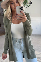 A woman wearing a long cardigan with pockets, paired with a white top and blue jeans, standing in front of a white brick wall.