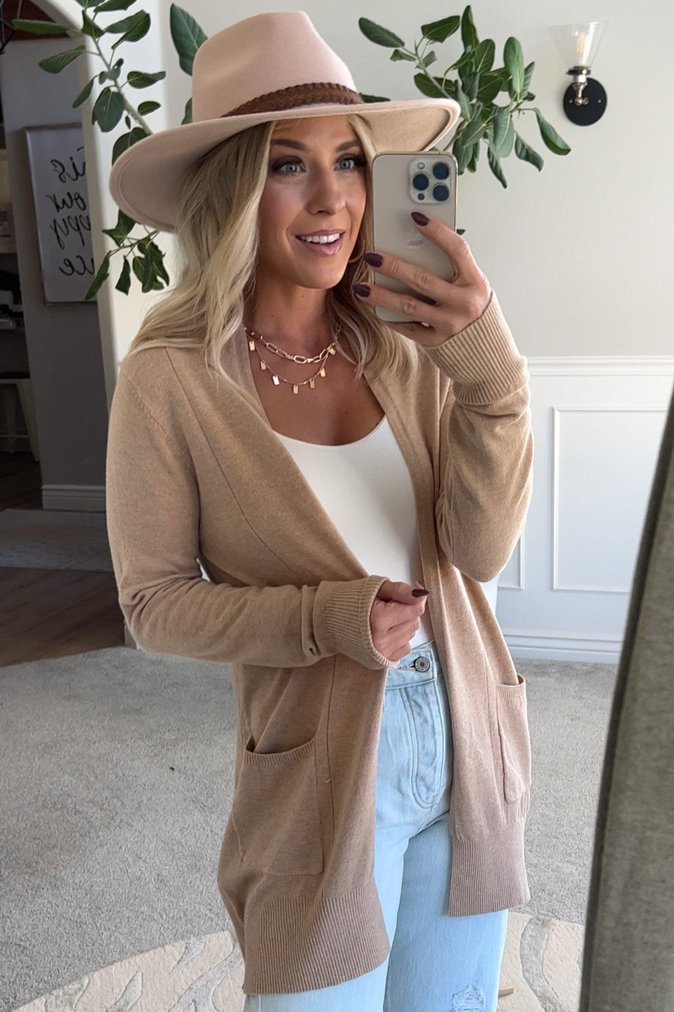 A woman wearing a long cardigan with pockets, paired with a white top and blue jeans, standing in front of a mirror.