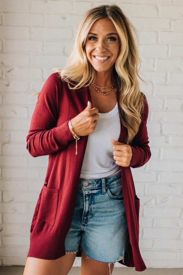 Woman wearing a red cardigan, white shirt, and denim shorts against a white brick wall.