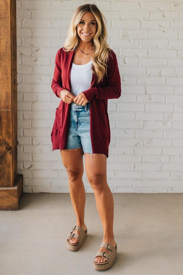 Woman wearing a burgundy cardigan, white top, denim shorts, and sandals against a white brick wall.