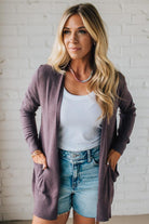 Woman wearing a purple cardigan over a white top and denim shorts against a white brick wall.
