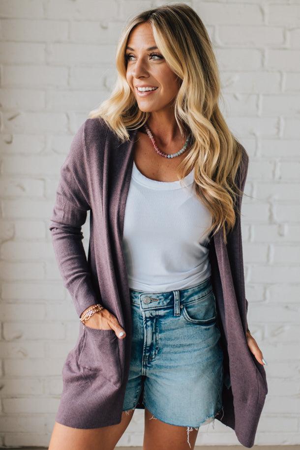 Woman wearing a purple cardigan over a white top and denim shorts against a white brick wall.