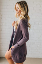 Woman wearing a long, dark purple cardigan against a white brick wall.