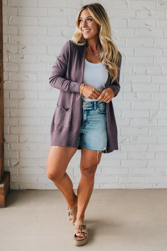Woman wearing a purple cardigan, white top, and denim shorts against a white brick wall.