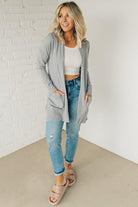 A woman wearing a long cardigan with pockets, paired with a white top and blue jeans, standing in front of a white brick wall.