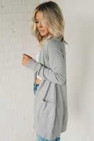 A woman wearing a long cardigan with pockets, paired with a white top and blue jeans, standing in front of a white brick wall.