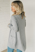 A woman wearing a long cardigan with pockets, paired with a white top and blue jeans, standing in front of a white brick wall.a