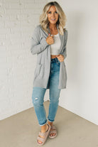 A woman wearing a long cardigan with pockets, paired with a white top and blue jeans, standing in front of a white brick wall.