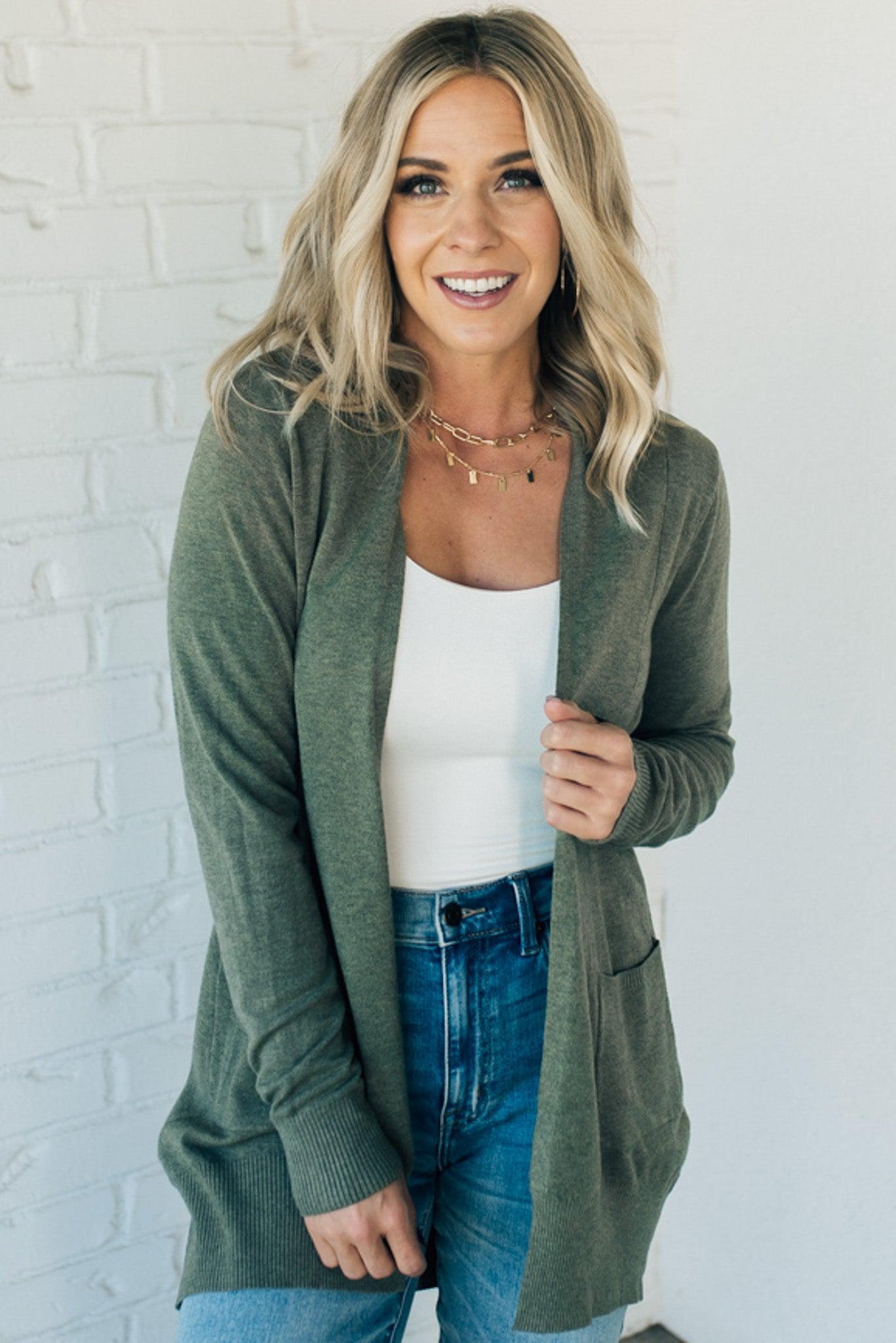 A woman wearing a long cardigan with pockets, paired with a white top and blue jeans, standing in front of a white brick wall.