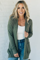 A woman wearing a long cardigan with pockets, paired with a white top and blue jeans, standing in front of a white brick wall.
