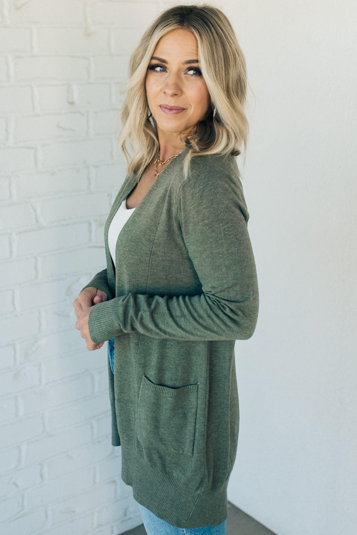 A woman wearing a long cardigan with pockets, paired with a white top and blue jeans, standing in front of a white brick wall.