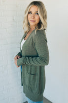 A woman wearing a long cardigan with pockets, paired with a white top and blue jeans, standing in front of a white brick wall.