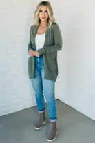 A woman wearing a long cardigan with pockets, paired with a white top and blue jeans, standing in front of a white brick wall.