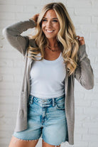 Woman wearing a white tank top, gray cardigan, and denim shorts against a white brick wall.