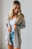 Woman wearing a gray cardigan over a white top and denim shorts against a white brick wall.