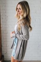Woman wearing a gray cardigan over a white top and denim shorts against a white brick wall.