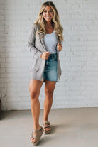 Woman wearing a gray cardigan, white tank top, and denim shorts against a white brick wall.