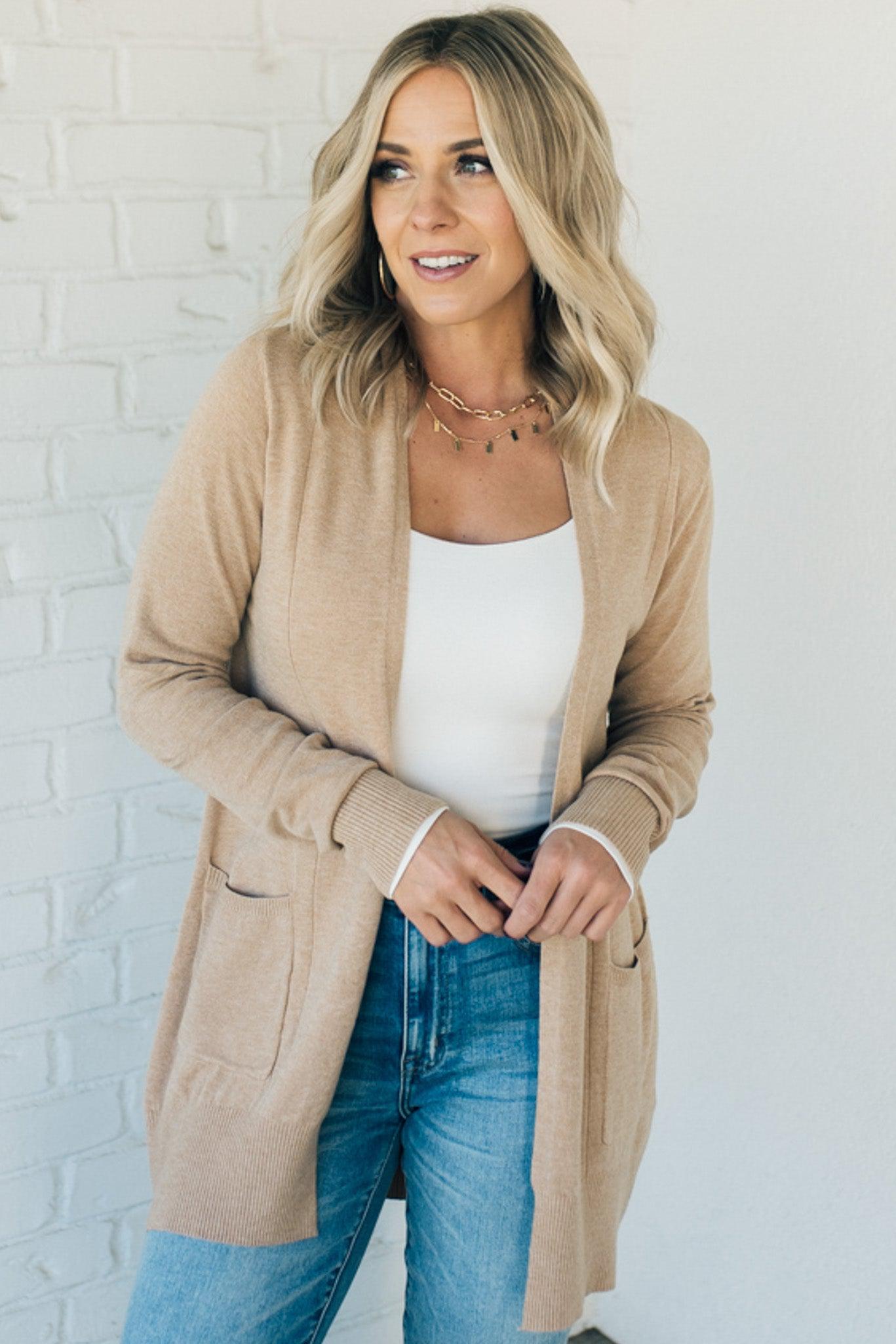 A woman wearing a long cardigan with pockets, paired with a white top and blue jeans, standing in front of a white brick wall.