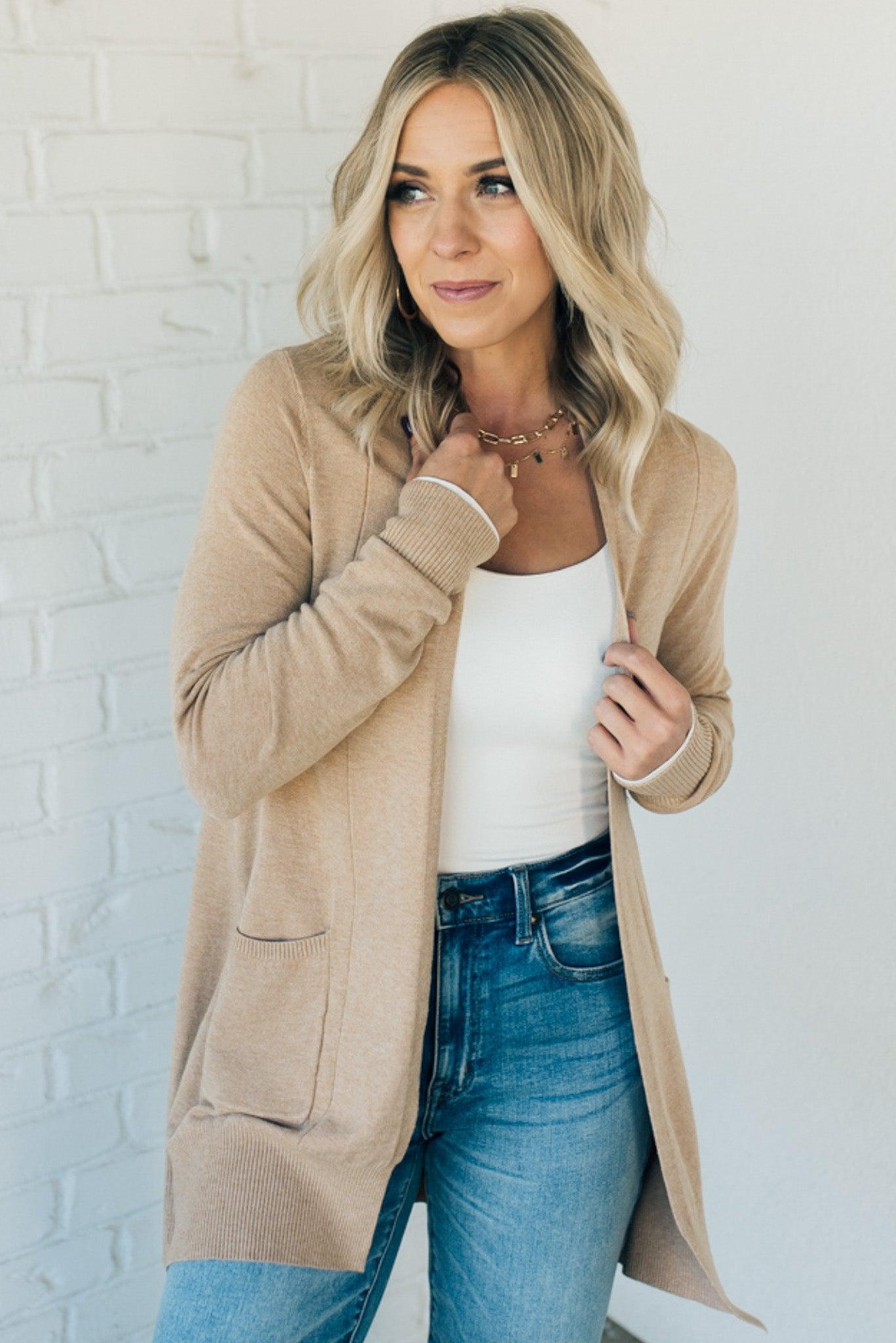 A woman wearing a long cardigan with pockets, paired with a white top and blue jeans, standing in front of a white brick wall.