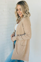 A woman wearing a long cardigan with pockets, paired with a white top and blue jeans, standing in front of a white brick wall.