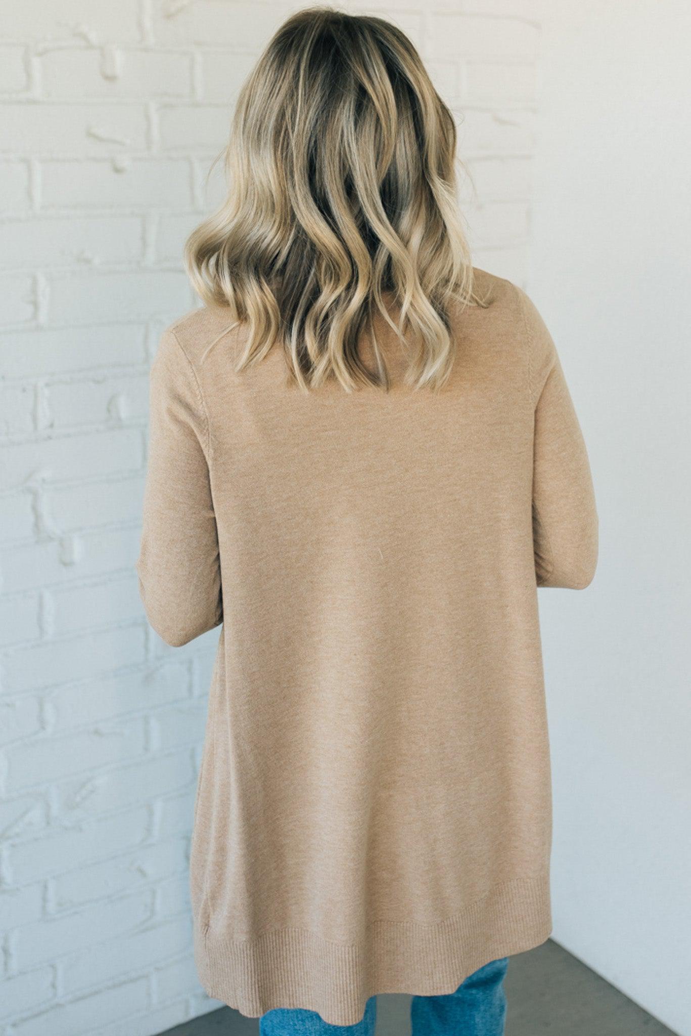 A woman wearing a long cardigan with pockets, paired with a white top and blue jeans, standing in front of a white brick wall.