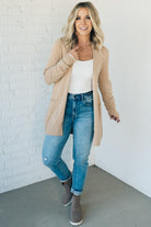 A woman wearing a long cardigan with pockets, paired with a white top and blue jeans, standing in front of a white brick wall.