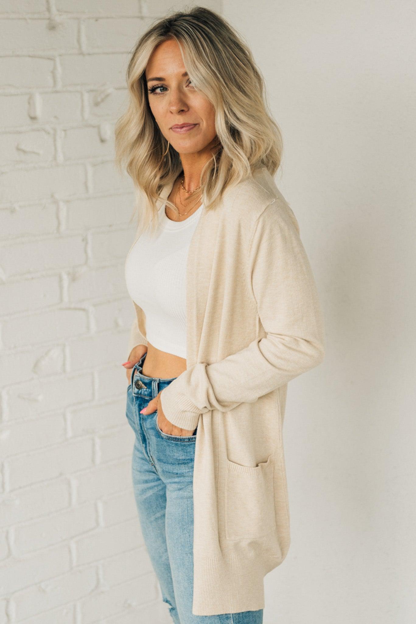 A woman wearing a long cardigan with pockets, paired with a white top and blue jeans, standing in front of a white brick wall.