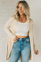 A woman wearing a long cardigan with pockets, paired with a white top and blue jeans, standing in front of a white brick wall.